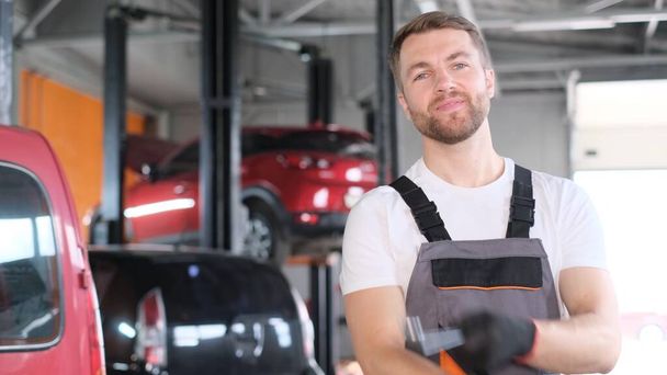 Retrato de un atractivo mecánico sonriente de aspecto americano de pie sobre el fondo de un taller de automóviles y sosteniendo una llave de coche. empleado feliz - Foto, Imagen
