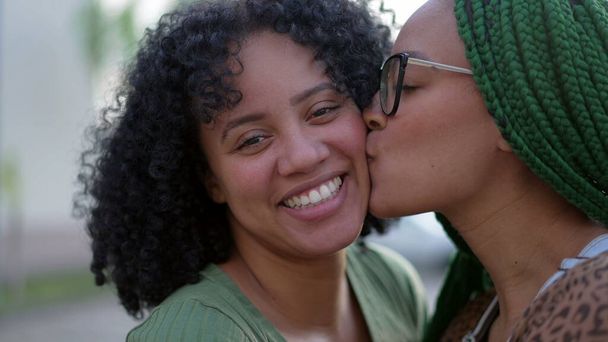 Two black sisters showing love and affection. African American sibling kissing friend in cheek. Happy Hispanic South American people care - Photo, Image