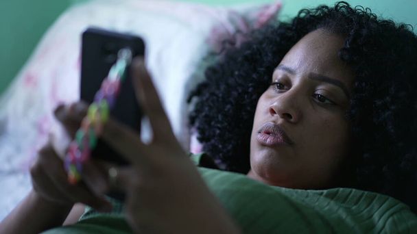 Eine junge schwarze Frau liegt im Bett und schaut auf ihr Mobiltelefon - Foto, Bild