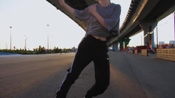 Sıska, açık renk saçlı, bol tişörtlü, gün batımında köprünün altında dans eden güzel bir kadın. Orta çekim - Video, Çekim