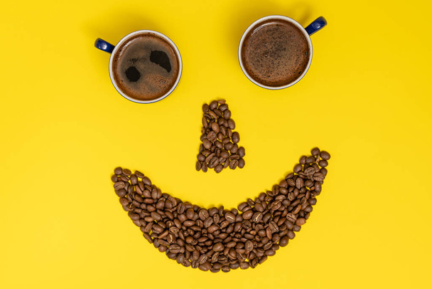 Mosolygó emotikon, kávébabból készült ajkak és kávéscsészékből készült szemek, sárga alapon történő kompozíció. - Fotó, kép