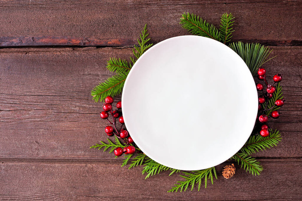Christmas or winter table setting mock up. Blank white plate with frame of evergreen branches and red berries. Top view on a dark wood background. Copy space. - Valokuva, kuva