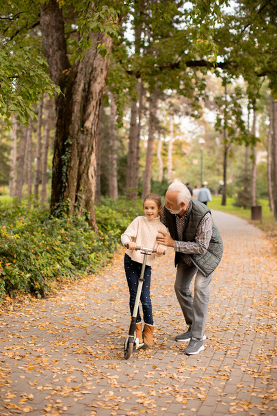 Hombre mayor enseñando a su nieta a montar patinete scooter en el parque de otoño - Foto, Imagen