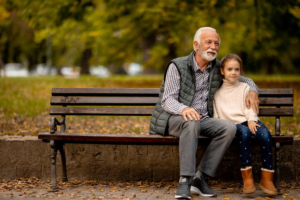 Handsome grandfather spending time with his granddaughter on bench in park on autumn day - Fotografie, Obrázek
