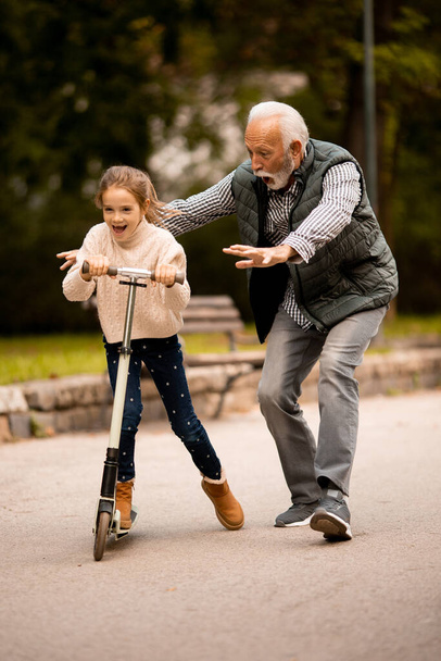 Старший мужчина учит свою внучку кататься на скутере в осеннем парке - Фото, изображение