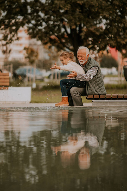 Handsome grandfather spending time with his granddaughter by small water pool in park on autumn day - Foto, imagen