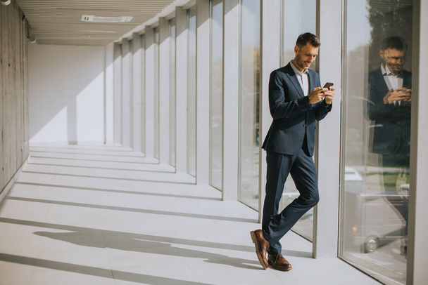 Bonito jovem homem de negócios usando telefone celular no corredor de escritório moderno - Foto, Imagem