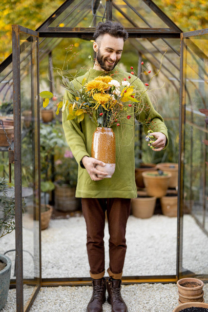 Πορτρέτο ενός ανθοπώλη άνθρωπος που στέκεται με ένα όμορφο μπουκέτο λουλούδια μπροστά από το μικροσκοπικό θερμοκήπιο στον κήπο. Έννοια του εργαστηρίου ανθοκομίας και λουλουδιών - Φωτογραφία, εικόνα
