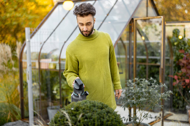 Stylish guy takes care of plants spraying on on them in garden with glasshouse on background. Gardening hobby and topiary plants concept - Valokuva, kuva