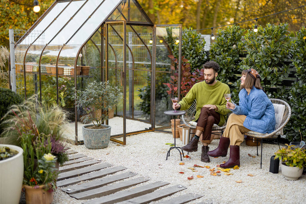 Young stylish couple hang out together, sitting with wine by the fire, spending evening time at cozy atmosphere in beautiful backyard with greenhouse on background - Zdjęcie, obraz