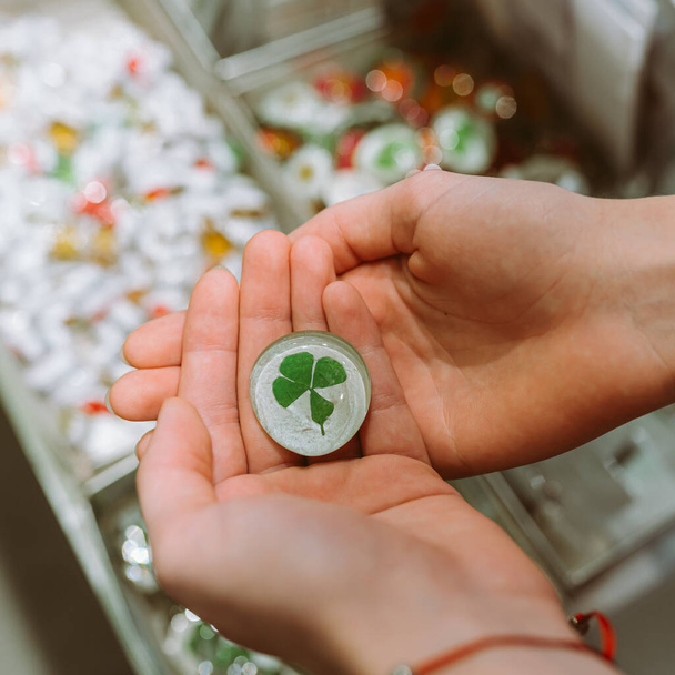 four-leaf clover in keychain in hands of girl, talisman of good luck in all endeavors. woman holds green four-leaf clover in hands, enclosed in keychain for memory and good luck, top view - Photo, Image