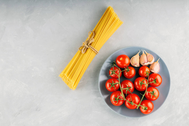 Nudelspaghetti mit Tomaten und Knoblauch auf grauem Hintergrund. Ein Bündel Spaghetti mit Bindfäden und Zutaten für italienische Pasta. Ansicht von oben. Kopierraum - Foto, Bild