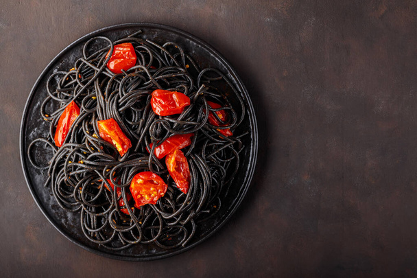 Черные спагетти с помидорами черри на черной тарелке. Вареные черные спагетти с красными помидорами на тёмном фоне. Вид сверху. Копирование пространства - Фото, изображение