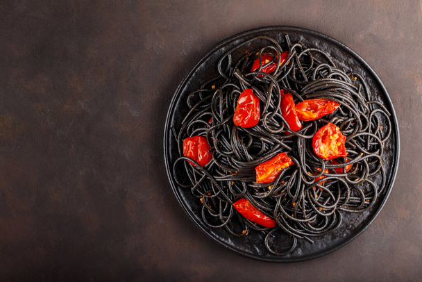 Black spaghetti with cherry tomatoes on a black plate. Boiled black spaghetti pasta with red tomatoes on dark background. Top view. Copy space - Photo, image