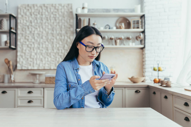 Young beautiful Asian woman sitting in kitchen at home and using mobile phone. Scrolls through the news feed, sits on social networks, checks mail. - Fotografie, Obrázek