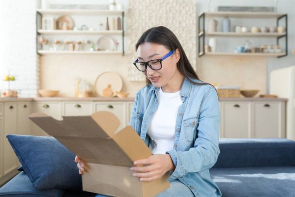 A young beautiful Asian woman received a package by a courier with home delivery. Sitting on the sofa at home and opening the box, online order from the store. - Valokuva, kuva