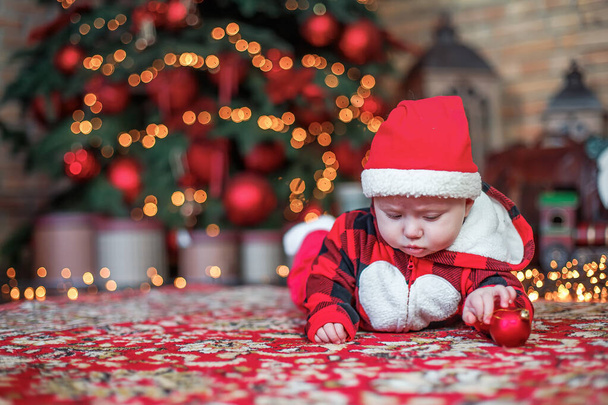 Маленький шестимесячный ребенок, одетый как Санта Клаус. Предыстория рождественской открытки. ребенок смотрит вниз на место для надписи на фоне светящихся гирлянд и рождественской елки. Копирование пространства - Фото, изображение