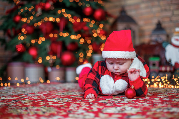 Ένα μωρό έξι μηνών ντυμένο Άγιος Βασίλης. Ιστορικό για χριστουγεννιάτικη κάρτα. παιδί κοιτάζει κάτω στη θέση για την επιγραφή στο φόντο των φωτεινών γιρλάντες και χριστουγεννιάτικο δέντρο. Αντιγραφή χώρου - Φωτογραφία, εικόνα