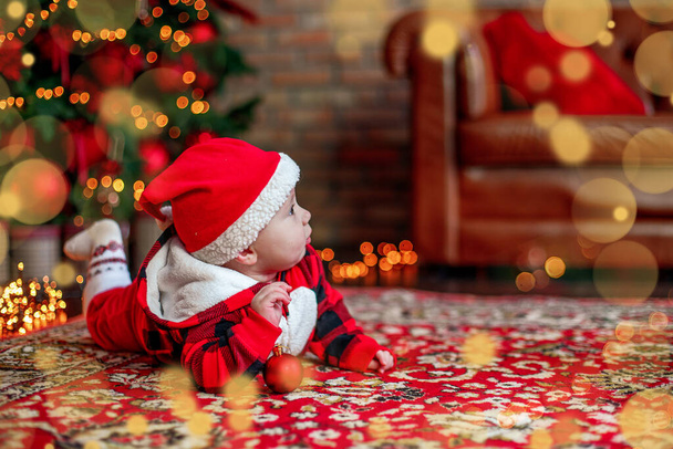 Little six month old baby dressed as Santa Claus. Background for christmas card. The child looks up at the place for inscription on background of luminous garlands and a Christmas tree. Copy space - Photo, Image