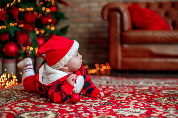 Kleines sechs Monate altes Baby als Weihnachtsmann verkleidet. Hintergrund für Weihnachtskarte. Das Kind blickt zur Stelle auf, um auf dem Hintergrund leuchtender Girlanden und eines Weihnachtsbaums eine Inschrift zu finden. Kopierraum - Foto, Bild