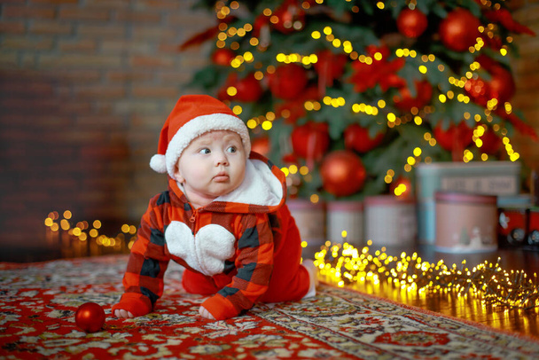 Kleines sechs Monate altes Baby als Weihnachtsmann verkleidet. Hintergrund für Weihnachtskarte. Kind blickt auf Platz für Inschrift auf Hintergrund von leuchtenden Girlanden und Weihnachtsbaum. Kopierraum - Foto, Bild
