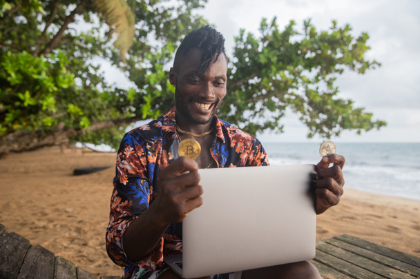 Africký obchodník s kryptoměn kontroluje trh se svým notebookem na pláži. - Fotografie, Obrázek