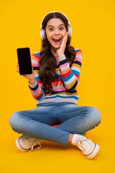 Teenager girl use smart phone, share social media, chatting by mobile phone wear stylish casual trendy clothes isolated over yellow background. Kid showing blank screen mobile phone, mockup copyspace - Fotografie, Obrázek
