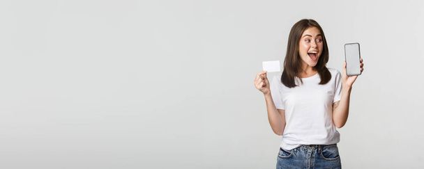 Ενθουσιασμένος και έκπληκτος χαριτωμένο κορίτσι δείχνει πιστωτική κάρτα και κινητό τηλέφωνο τραπεζική εφαρμογή στην οθόνη. - Φωτογραφία, εικόνα