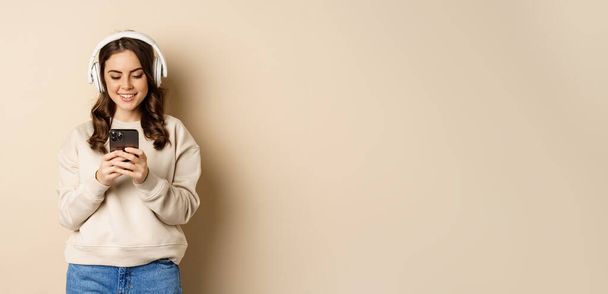 Smiling cute girl in headphones, looking at mobile phone, listening music or podcast, standing over beige background. - Foto, Imagem