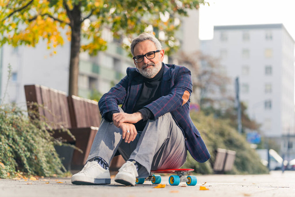 A smiling, elegant, bearded, grey-haired mature man with his glasses on, sitting on a red penny board in the city park, posing and looking at the camera. High quality photo - Foto, immagini