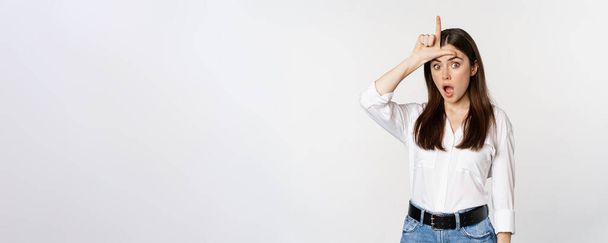 Young woman mocking loser, showing L word sign on forehead and gasping with disbelief surprise, standing against white background. - Foto, Imagem