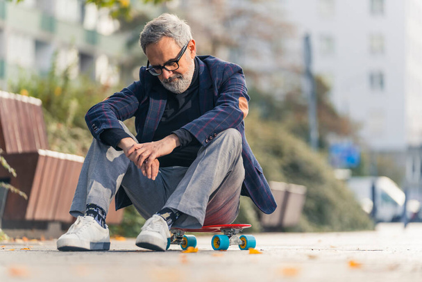 Elegante bebaarde grijsharige volwassen man met bril zittend op een skateboard in het stadspark. Hoge kwaliteit foto - Foto, afbeelding