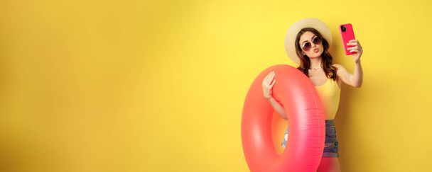 Stylish brunette girl on vacation, taking selfie with swim ring, going on beach, swimming in sea on summer holiday, standing over yellow background. - 写真・画像