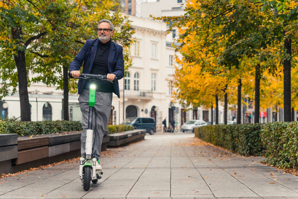 Millennial modern bearded grey-haired mature man with eyeglasses on driving an electro scooter in the park alley and enjoying warm weather during the fall season. High quality photo - Zdjęcie, obraz