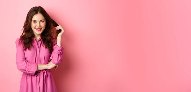 Beautiful brunette woman enjoying spring, wearing bright clothes and make up touching her curly hair and smiling confident at camera, standing against pink background. - Fotografie, Obrázek