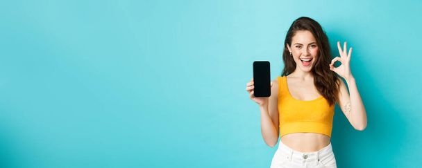 Για δες αυτό. Εμφανίσιμη γυναίκα με αυθάδης χαμόγελο, κλείσιμο του ματιού και εμφάνιση εντάξει σημάδι με άδεια οθόνη smartphone, συνιστώντας app, στέκεται πάνω από το μπλε φόντο. - Φωτογραφία, εικόνα