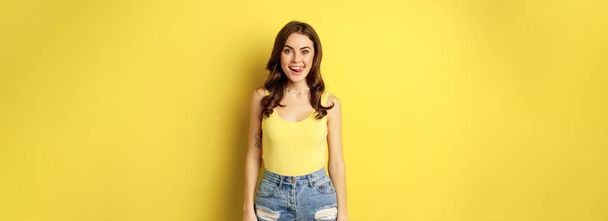 Stylish young brunette woman showing her white healthy teeth, smiling and showing tongue, winking enthusiastic, standing over yellow background. - Fotoğraf, Görsel