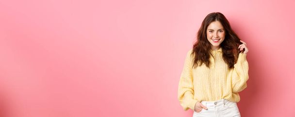 Красивая кокетливая девушка, играющая волосами, улыбающаяся и смотрящая на камеру, делая гламурную позу, стоя на розовом фоне. - Фото, изображение