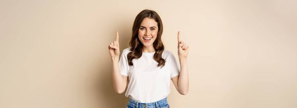 Enthusiastic young woman, female customer pointing fingers up and smiling, showing banner or logo, standing against beige background. - Фото, зображення