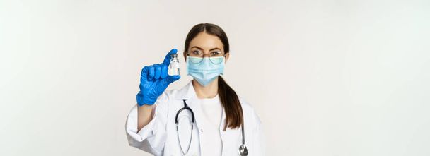 Portrait of young woman, doctor in medical face mask and uniform, showing vaccine, covid-19 vaccination campaign, standing over white background. - Foto, Imagem
