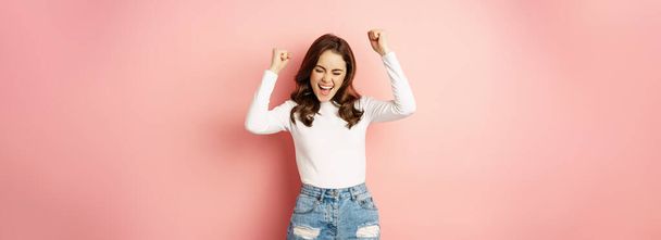 She is a winner. Enthusiastic brunette girl celebrating, jumping from happiness and shouting joyful, achieve goal success, standing over pink background. Victory concept - Фото, изображение