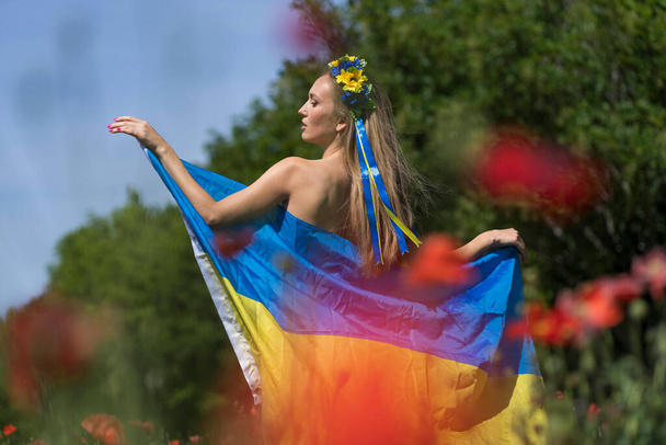 A young blonde Ukrainian woman stands in a field of Red Poppy flowers holding the flag of Ukraine showing her support for the war in her native country of Ukraine - Valokuva, kuva