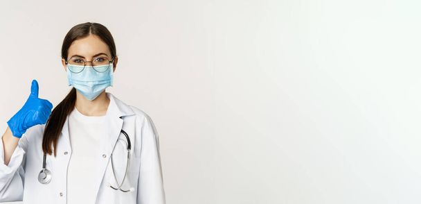 Close up portrait of woman doctor, physician in face mask from coronavirus, showing thumbs up and smiling, recommending clinic or medical website, white background. - Photo, Image