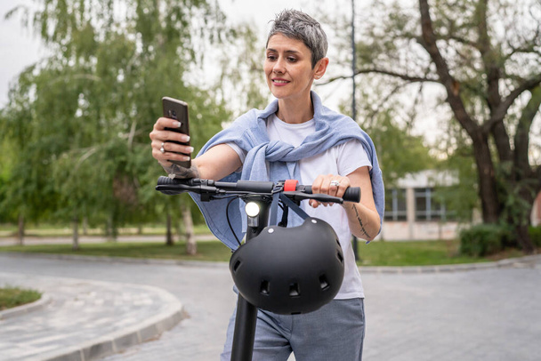 one woman mature senior caucasian female standing outdoor use mobile phone for selfie photos or video call by electric kick scooter in day confident modern alternative mode of transport copy space - Foto, Imagem