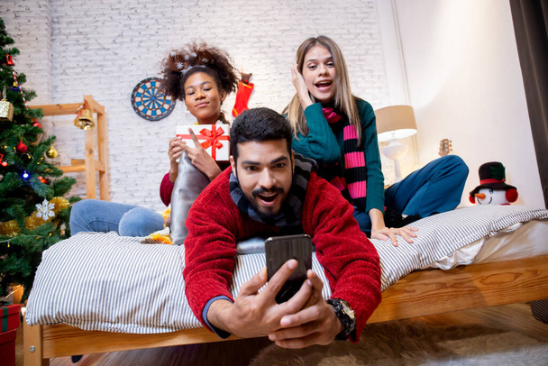 Grupa młodych przyjaciół robi selfie z podnieceniem i cieszyć się smartfonem razem w domu, mężczyzna i kobieta robiąc zdjęcie w Wigilię Bożego Narodzenia z wesołym, x-mas i nowy rok lub koncepcji wakacje. - Zdjęcie, obraz