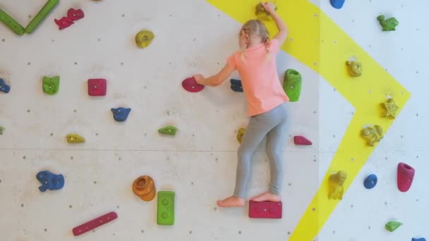 Brave child little blond girl is climbing up artificial wall in rock-climbing gym exercising. Childhood and extreme sports concept. - Footage, Video