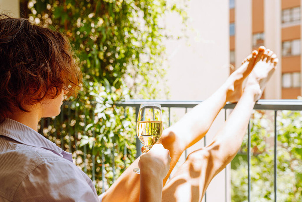 Schlanke Frau sitzt auf Balkon, mit einem Glas Wein oder Champagner, schlanke Beine auf Balkongeländer gestützt. In Sonnenstrahlen. Frau genießt Weißwein oder Champagner auf Balkon - Foto, Bild