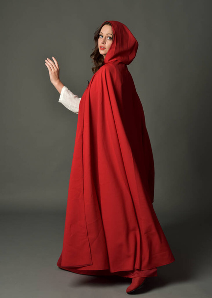 Full length portrait of woman wearing red medieval fantasy costume, flowing hooded cloak. Standing pose in backview, gestural hand poses, walking away from camera isolated on grey studio background. - Фото, изображение