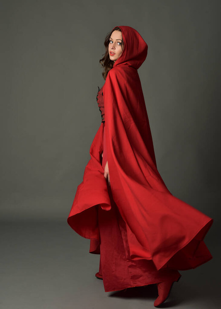 Volledig portret van een vrouw in rood middeleeuws fantasiekostuum, stromende capuchon mantel. Staande houding in achteraanzicht, gebaar hand poses, weglopen van camera geïsoleerd op grijze studio achtergrond. - Foto, afbeelding