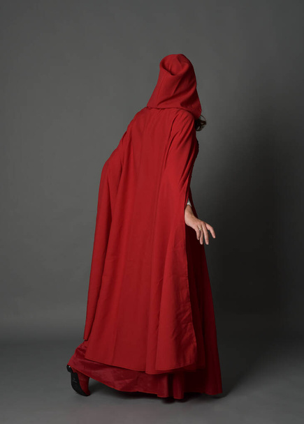 Full length portrait of woman wearing red medieval fantasy costume, flowing hooded cloak. Standing pose in backview, gestural hand poses, walking away from camera isolated on grey studio background. - Photo, Image